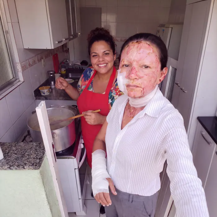 Ivone na cozinha, ao lado de uma mulher de avental vermelho que mexe uma panela grande